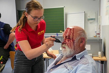 Einsatzkraft der DLRG beim Schminken einer weiteren Person mit einer größeren Verletzung am Kopf